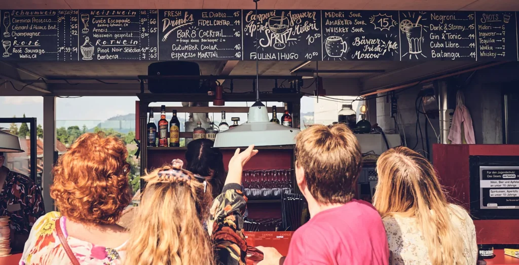 Mehrere Personen die vor einer Bar stehen und das Menu oberhalb der Bar betrachten.