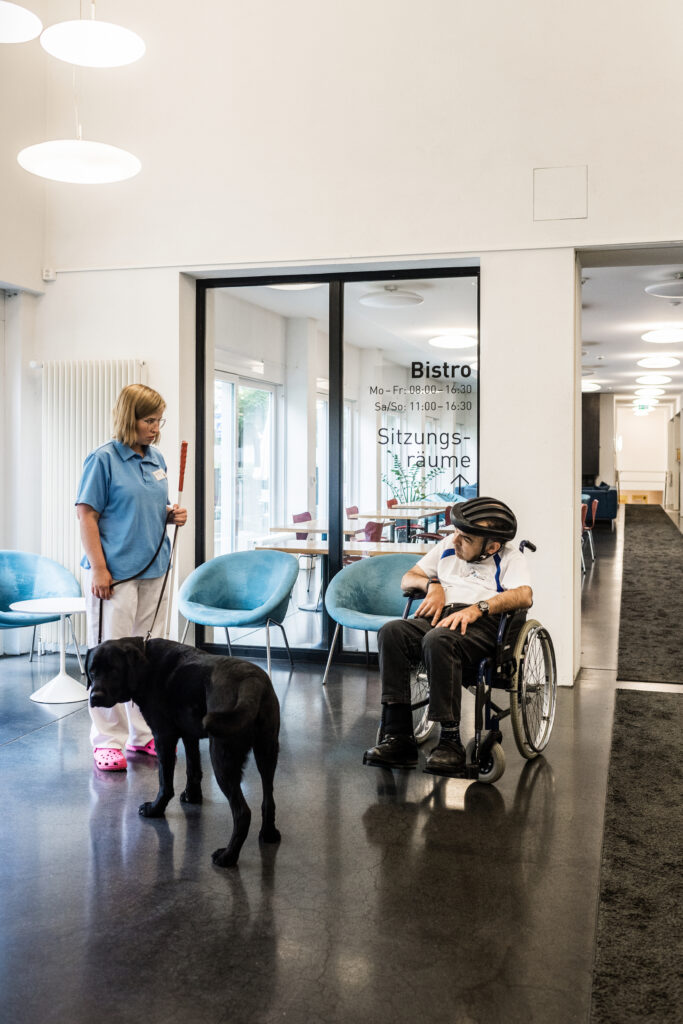 Pflegerin mit einem Blindenhund und einem Patienten im Rollstuhl.