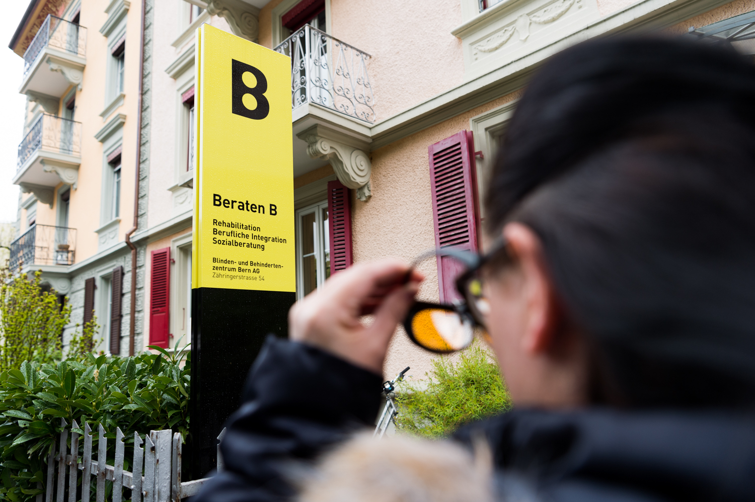 Person die vor einem Gebäude steht und die Brille von der Nase nimmt.
