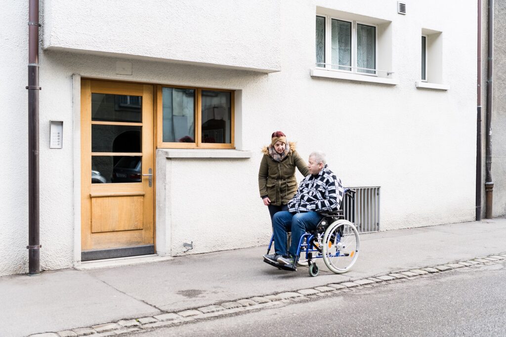 Junge Frau die draussen neben einem älteren Mann im Rollstuhl steht.