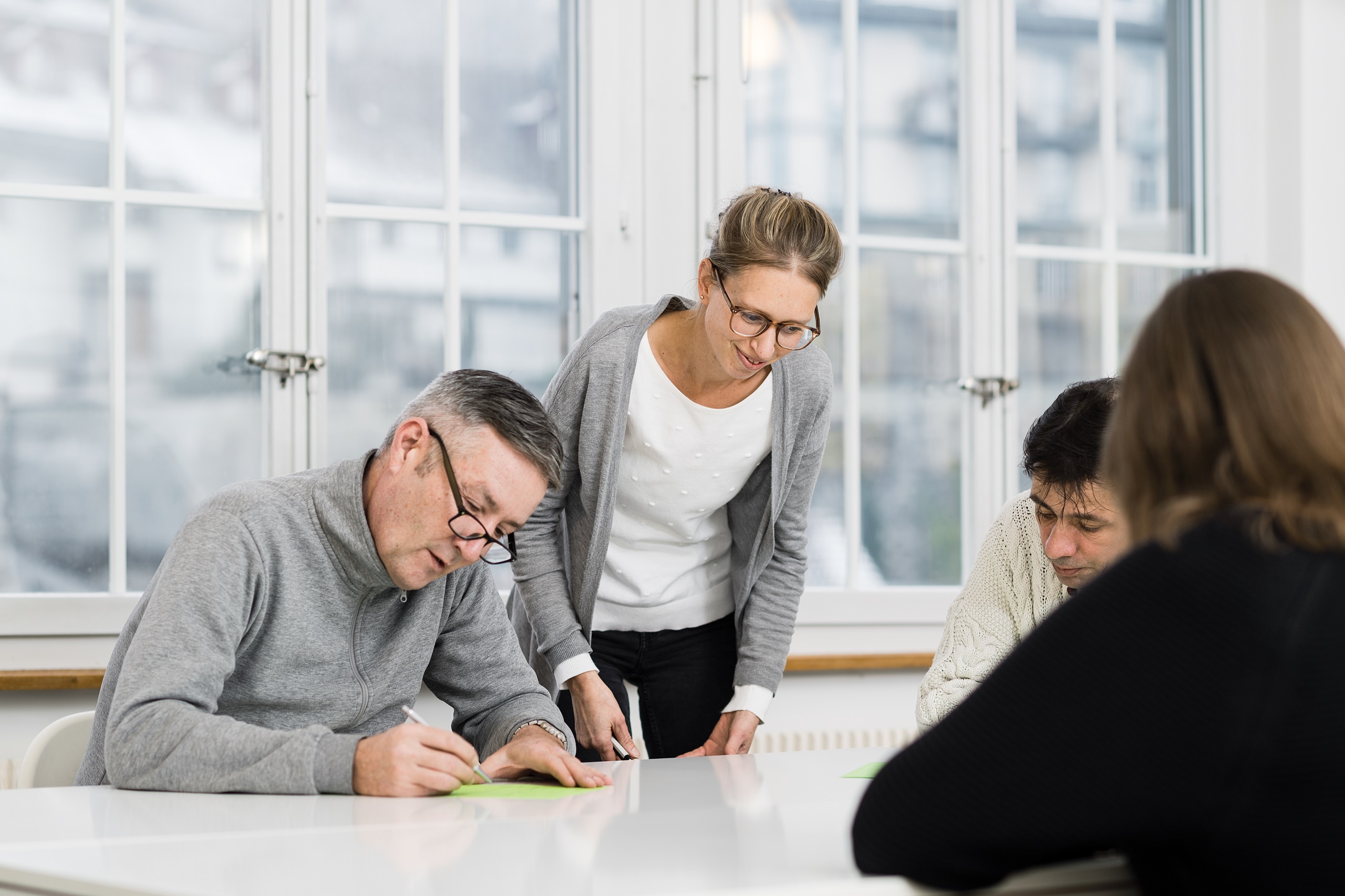 Personen die an einem Tisch sitzen und von einer Lehrerin etwas gezeigt bekommen.