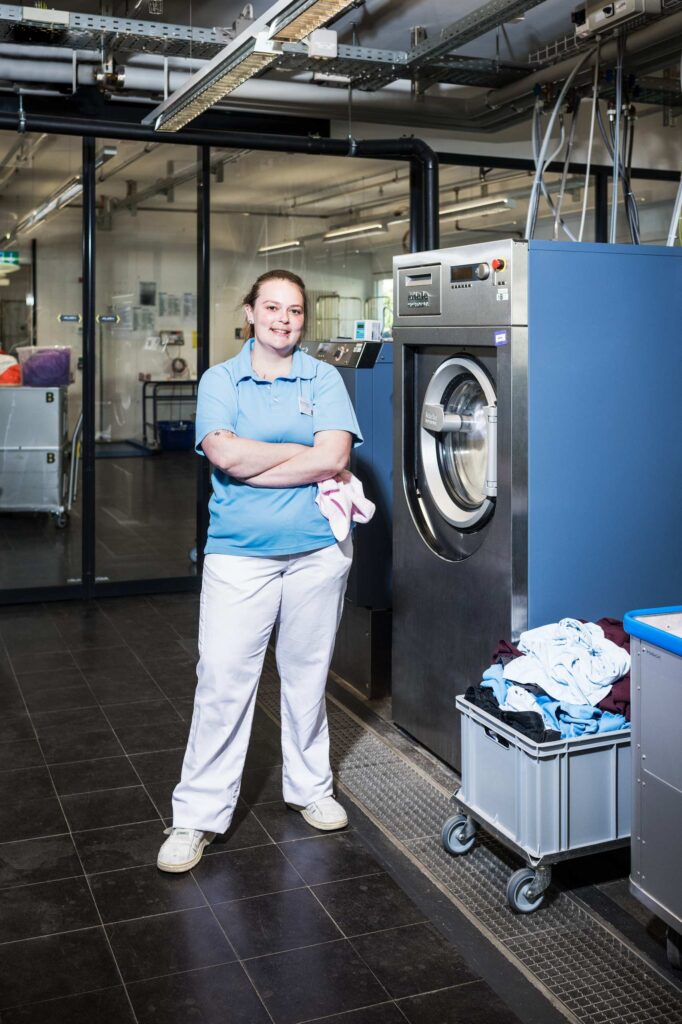 Bild zeigt, eine Mitarbeiterin vom B die in der Wäscherei des Betriebs arbeitet und in die Kamera lacht. 