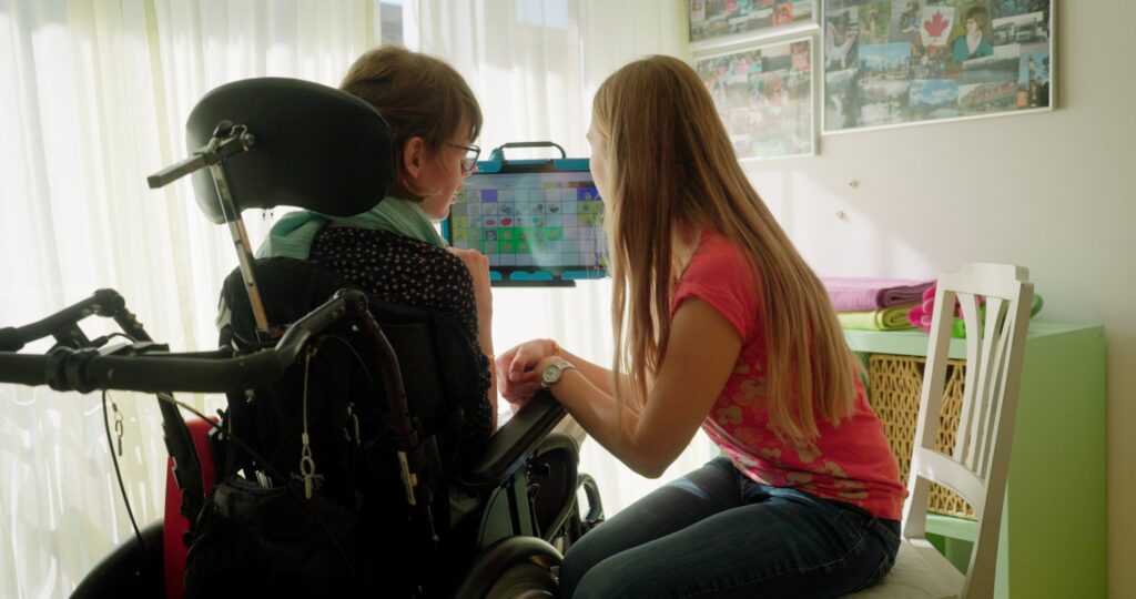 Eine junge Frau sitzt im Rollstuhl. Daneben sitzt eine zweite Frau. Beide schauen auf einen Bildschirm wo Symbole für unterstützte Kommunikation zu sehen sind.