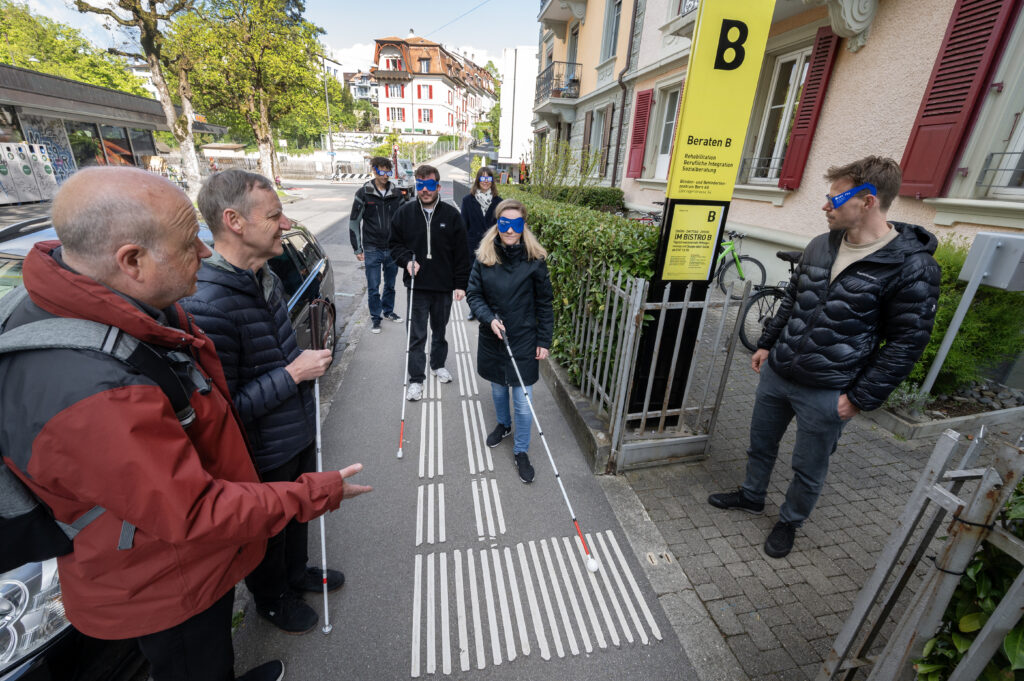 Teilnehmer des Workshops tragen eine Dunkelbrille und gehen mit Langstock den Leitlinien entlang.