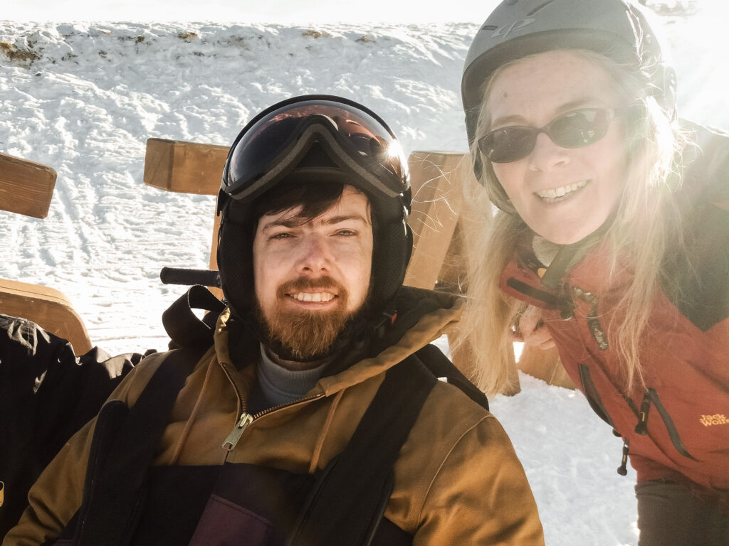 Zwei Personen im Schnee und Skihelm die in die Kamera lachen.