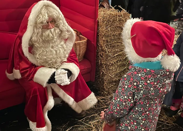 Kind mit roter Zipfelmütze sagt einen Vers auf vor dem Weihnachtsmann, der auf einem roten Sofa sitzt.