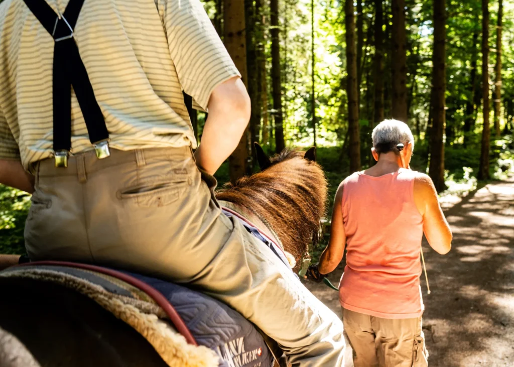 Bewohner auf einem Pferd, welches von einer Frau durch den Wald geführt wird.
