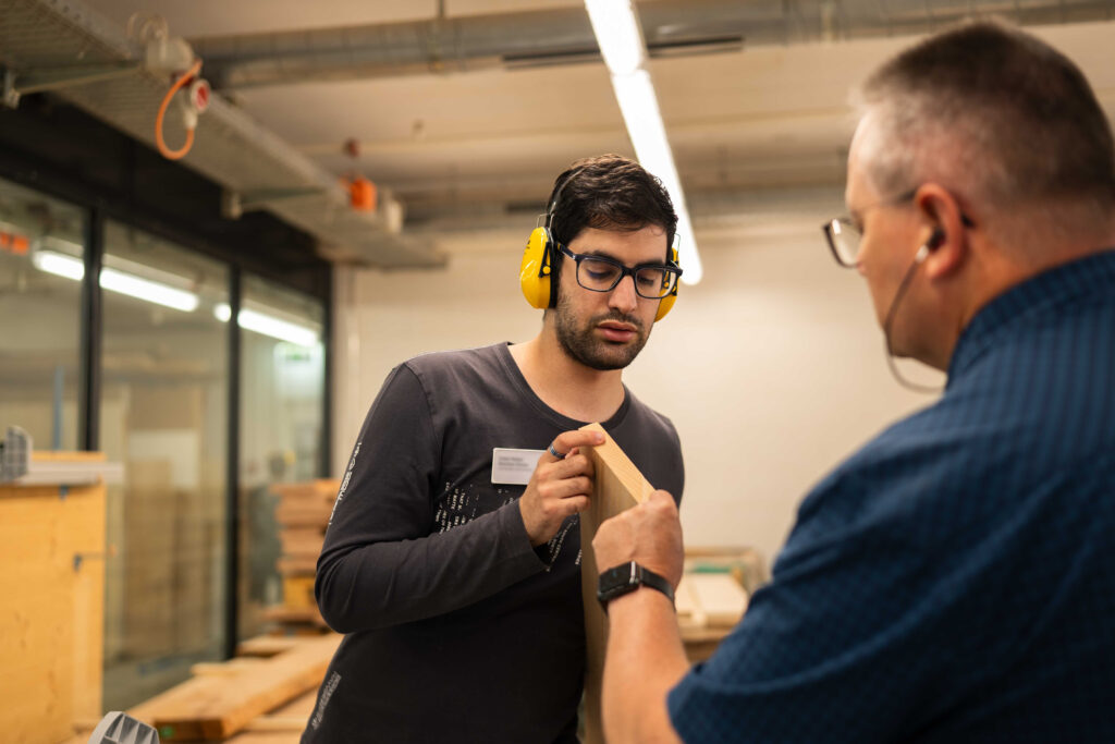 Zwei Männer in einer Werkstatt begutachten gemeinsam ein Holzstück; einer trägt Gehörschutz und erklärt konzentriert Details, während der andere aufmerksam zuhört.