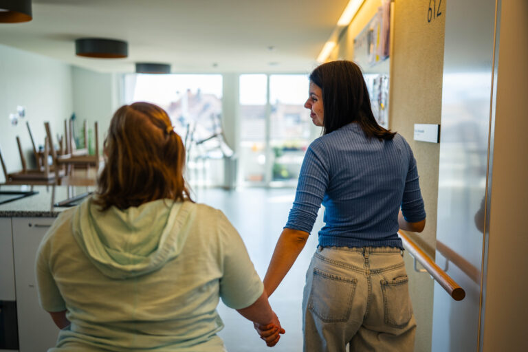 Zwei Frauen gehen Hand in Hand durch einen hellen Flur, während eine der beiden sich umdreht und zur anderen blickt.