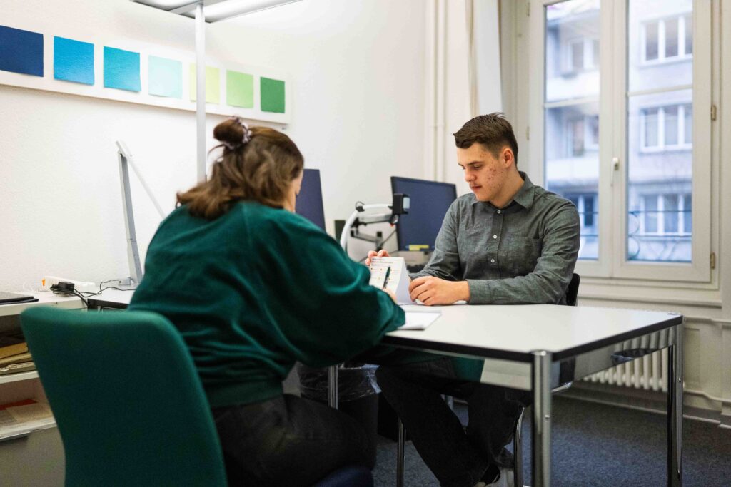 Zwei Personen sitzen sich in einem Büro an einem Tisch gegenüber und besprechen gemeinsam ein Dokument, während im Hintergrund Computerarbeitsplätze und ein Fenster zu sehen sind.